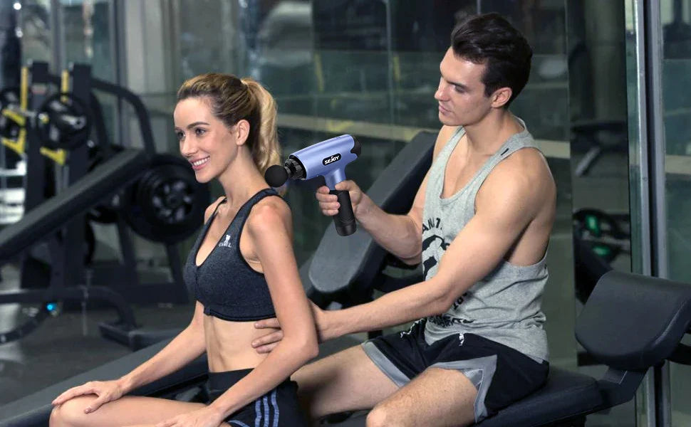 Man using Sejoy Fascia Gun massager on woman's back in gym setting.